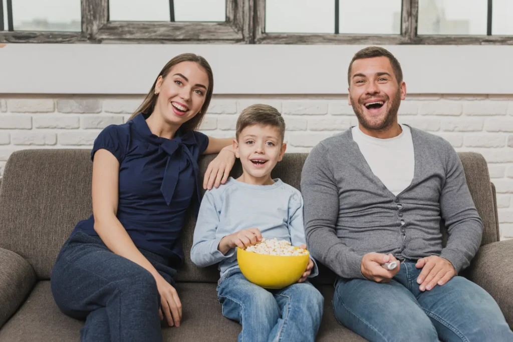 Famille souriante regardant la télévision ensemble grâce à leur abonnement IPTV 12 mois — parents et enfant sur le canapé avec du popcorn